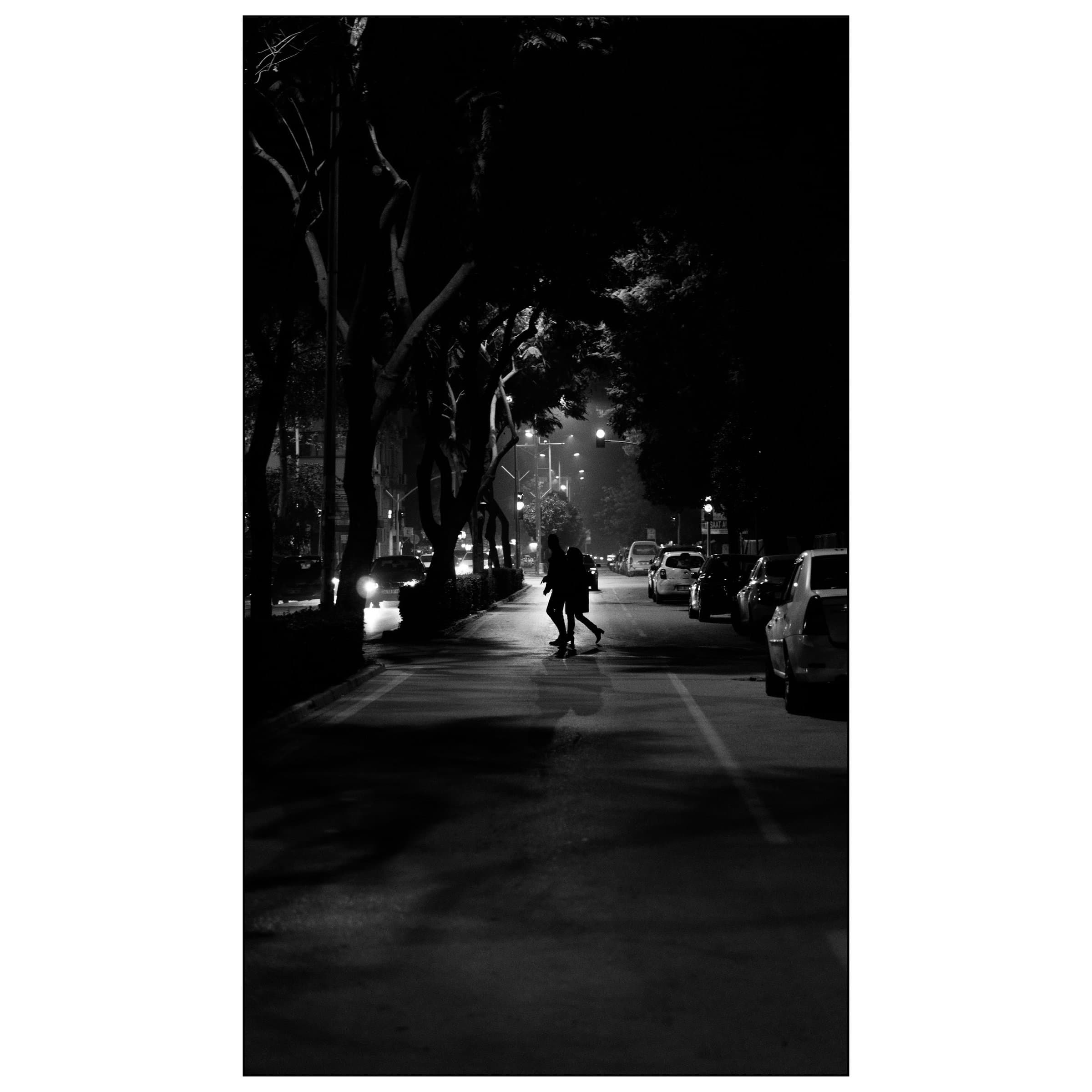 Gece ağaçlı cadde üzerinde araba ışıkları arasında yürüyen iki kişinin silueti