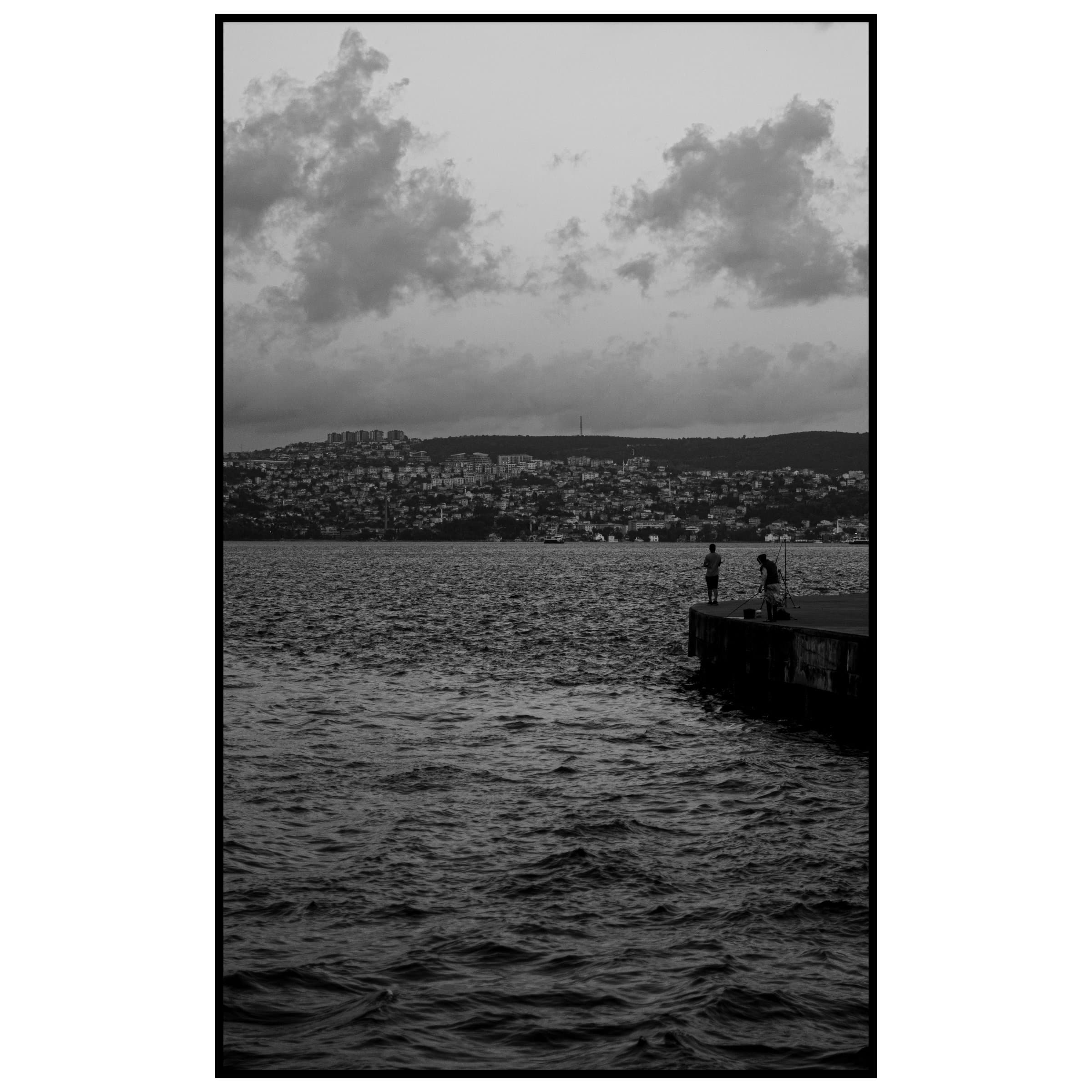 Deniz kenarındaki iskelede duran balıkçıların silueti ve karşı kıyıdaki şehir tepesi